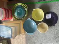 Top-down view of a selection of assorted plastic kitchen storage containers and mixing bowls in various colors and sizes arranged on the floor.
