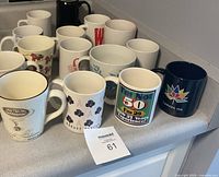 Overview photo showing the assortment of 16 coffee mugs arranged in three rows on a white counter, showcasing a variety of colors and designs including Tim Hortons, Canada 150 logo, and several patterned mugs.