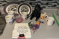 Photo of set showing 5 cat-themed mugs, 2 cat-illustrated plates, black ceramic cat figurine, fabric towel with cat pun, and 3 cat toys (balls and feather toys) arranged on kitchen counter.