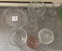 Nine decorative glass pieces displayed on carpeted surface. Included are a clear scalloped plate, a clear vase, multiple clear glass bowls of varying sizes, and a small pink handled glass dish.