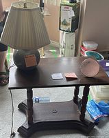 Table lamp with pleated beige shade and grey ceramic base placed on a dark wooden side table with turned legs and a lower shelf. Minor wear visible on table surface.