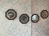 Four decorative wall plates hung on floral wallpapered wall, each in an octagonal dark wood frame, featuring vintage painted scenes behind glass.