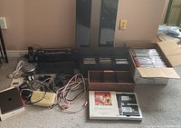 Overview of stereo, speakers, keyboard, telephone, box of CDs/DVDs, wooden CD organizer, VHS set, and iPad on carpeted floor.