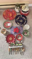 Overview of Christmas plates, serving dishes, butter spreaders, snow globe, and holiday container on carpeted floor.