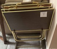 Four foldable TV tray tables stacked in the matching gold-tone metal stand showing front view with handle details on top edges.