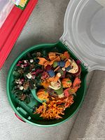 Green plastic bin opened showing assorted Halloween and autumn decorations including pumpkins, scarecrows, pine cones, and ribbons