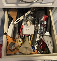 Drawer containing assorted kitchen utensils including spoons, spatulas, peelers, can opener, funnel, masher and other tools in mixed materials and colors.