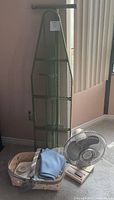 Full view of the set including the metal mesh ironing board, beige and white table fan, and woven basket with fabric and other contents inside.