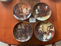 Four ceramic plates arranged on wooden table, featuring various Norman Rockwell inspired designs with human figures in vintage scenes.