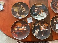 Four Norman Rockwell collector plates showing different family scenes arranged on a wooden table.
