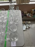 Top view showing 12 clear wine glasses arranged on a table with stems visible, along with nearby cups and boxes in background.