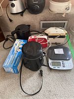 Various small kitchen appliances placed together on floor including blender, food steamers, hand mixer, digital kitchen scale, electric rice cooker, electric cutting utensil set and sandwich grill machine.