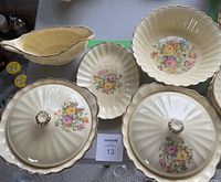 Photo showing six creamy yellow ceramic serving pieces with fluted edges, floral cross-stitch-like designs in the centers, and 18k gold trim. Pieces include covered dishes with gold-accented knobs, a gravy boat, a large bowl, and a platter.