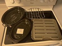 Two large black speckled oval roasting pans with lids, a metal roasting rack, and a rectangular grilling tray with ventilation slots placed on stove top.