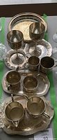 Silver-plated vintage goblets, trays, cups, and serving items arranged on a table. Items show signs of age and patina consistent with vintage condition.