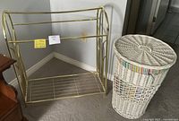 White brass quilt rack and white wicker laundry basket with colorful fabric lining and lid shown side by side on carpeted floor.