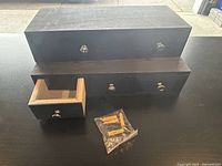 Two black wooden wall-mounted mini drawer organizers, one with an open drawer and mounting hardware visible in a plastic bag.