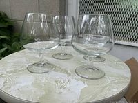Four clear glass brandy snifters arranged on a round table outdoors, showing their rounded bowl design and clear transparency.