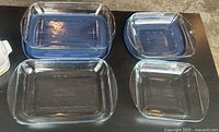 Two rectangular glass dishes, one with and one without blue lid, alongside two square glass dishes, one with and one without blue lid, all clear tempered glass.