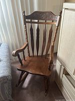 Front angle of antique wooden rocking chair showing carved top backrest, vertical slats, contoured seat, and curved armrests with turned supports.
