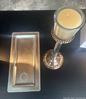 Birds-eye view of a tall metal and glass candle holder with a cream pillar candle inside and a rectangular silver-tone metal trinket tray with beaded edge on a dark surface, showing slight wear on the tray.