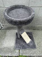 Grey carpeted small cat tree with round cushioned top perch and angled sisal-wrapped scratching post on a square base, placed on paving stones against a grey block wall.