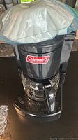 Front view of Coleman coffee maker covered with plastic bag, showing logo and glass carafe.