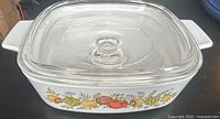 Square white ceramic CorningWare casserole dish with colorful vegetable illustrations on the sides and clear glass lid with knob handle, shown on a black surface.