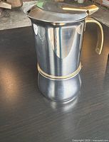 Side view of the stainless steel stovetop espresso maker on a dark surface showing brass handle and gold band accent, in vintage used condition.