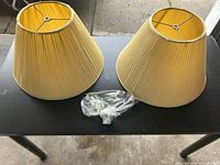 Two pleated cream beige lampshades set on a black table with original finials packaged in a plastic bag in front.