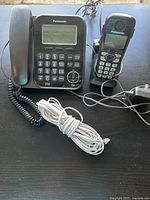 Panasonic black corded home phone with large numbered keypad and digital screen, accompanied by a black cordless handset on charging base, power adapter, and telephone cord.