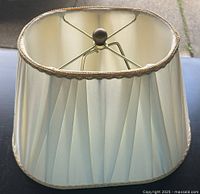 Top angled view of an oval ivory pleated lampshade with decorative gold trim and visible metal frame.