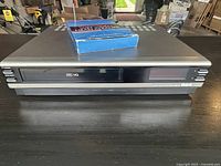 Black RCA VHS HQ VCR with visible front controls and display panel placed on dark surface.