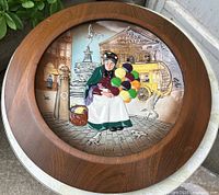 Frontal view of the round Royal Doulton ceramic character plate featuring a relief of an elderly woman holding colorful balloons in front of The London Pavilion, mounted in a circular wood display frame.