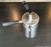 Photo of vintage stovetop espresso maker showing brushed aluminum body and black ribbed handle with locking lever lid in closed position from above.