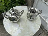 Coffee pot and creamer shown on a round marble-top table, plastic wrapped for protection, ornate scroll handles visible.