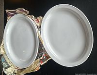 Two of the white ceramic oval platters stacked with another platter next to them on a table showing their smooth, glossy finish and oval shape with raised rims.