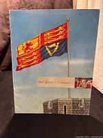 Front cover of the souvenir program showing three flags on a stone structure and the title 'Our Queen is Crowned'.