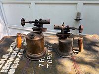 Two vintage antique brass blow torches with wooden handles, showing patina and wear, viewed from side angle on metal surface.