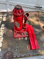 Front angle of red hydraulic bottle jack showing the top lifting saddle and two detached red handles on a metal surface with paint wear.
