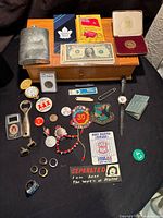 Full overview of the assorted small items including coins, pins, flask, wooden drawer, jewelry and other collectibles.