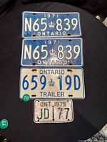 Four vintage Ontario licence plates laid on black fabric: two 1971 passenger plates (N65 839), one 1971 trailer plate (659 19D), one 1975 plate (JD 177).