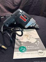 Photo of the vintage SKIL electric drill with visible power cord and included instruction manual on a black backdrop