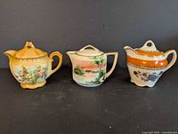 Front view of three single serve teapots with distinct floral and scenic decorations on a black background.