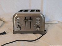 Photo of a stainless steel 4 slice toaster showing front panel with dual controls, multiple buttons for bagel, defrost, reheat and cancel, two browning knobs, and power cord.