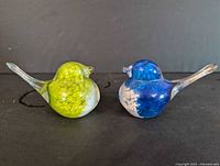Side view of two blown glass bird figurines, one green and white, one blue and white with clear glass features.