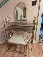 Vanity table with glass top, arched mirror, and cushioned stool.