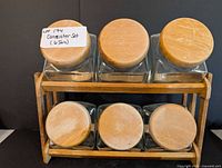 Six clear glass canisters with round wooden lids arranged on a wooden rack, viewed from front and slightly above, showing the tops of the lids with visible natural wood texture and some signs of wear.
