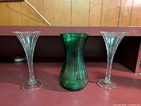 Three glass vases on a shelf: one green with ribbed vertical lines and two clear glass with hexagonal bases and flared tops.