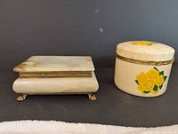 Two closed jewelry boxes, one rectangular marble with brass feet and one round porcelain with yellow rose decoration